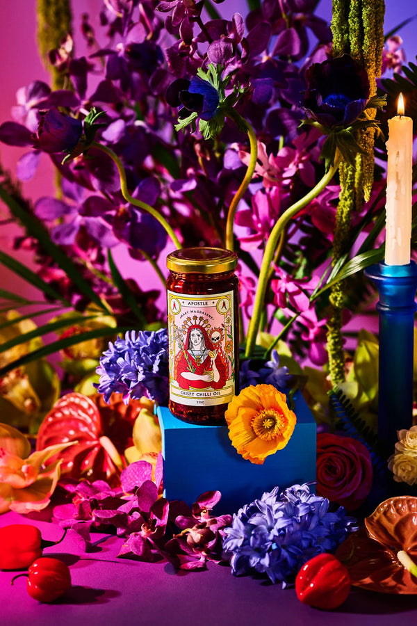 Apostle Crispy Chilli Oil jar styled among vibrant purple, orange, and red flowers with a lit candle, creating a bold and colourful display scene