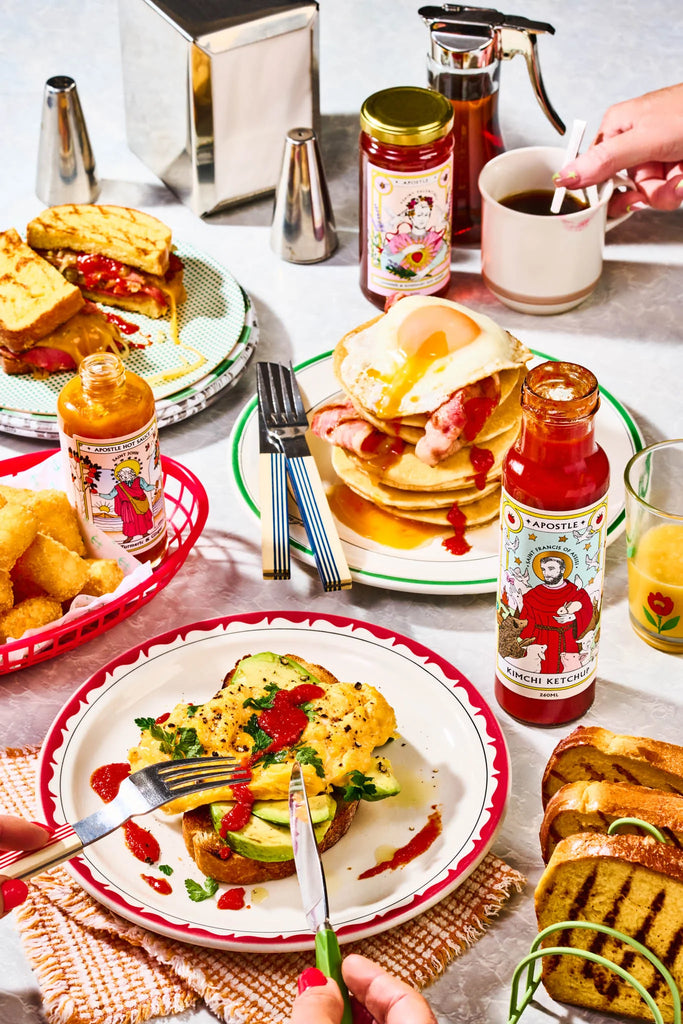 Vibrant brunch spread featuring scrambled eggs on toast, pancakes with bacon and egg, and toasted sandwiches, all topped with Apostle Kimchi Ketchup and hot sauces