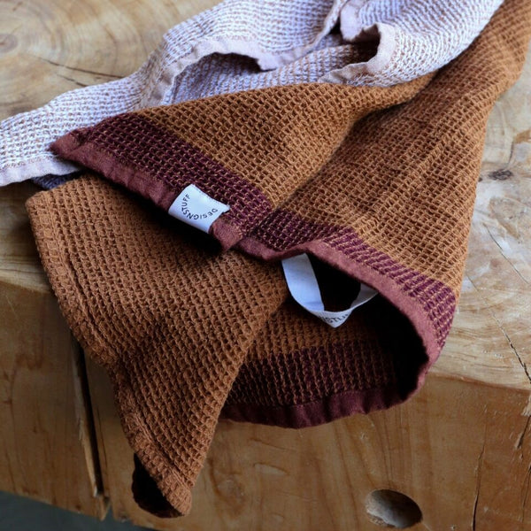 A close-up of two waffle weave tea towels in warm brown and burgundy hues, one showing a visible “Designstuff” tag, styled casually on a rustic wooden surface.