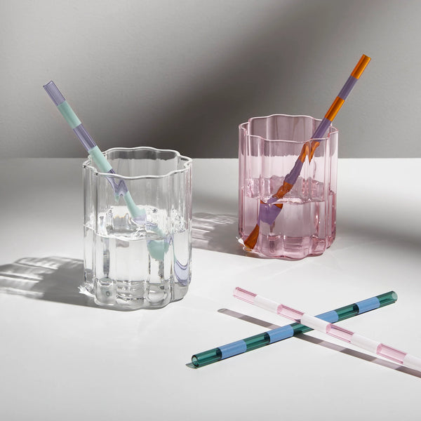 Striped glass straws in pastel and jewel tones styled in pink and clear scalloped glasses, with two straws placed diagonally on a white surface.