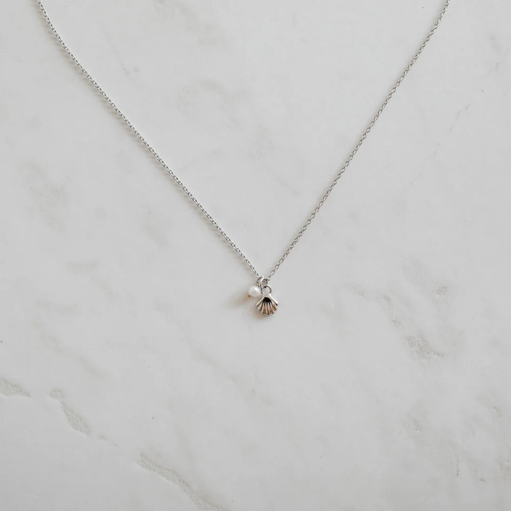 Minimalist silver necklace featuring a dainty shell charm and pearl pendant, styled flat on a light marble surface for an elegant, ocean-inspired look.