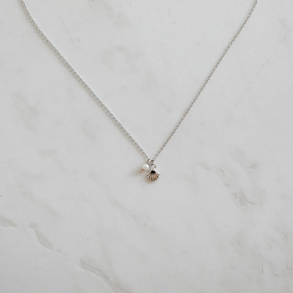 Minimalist silver necklace featuring a dainty shell charm and pearl pendant, styled flat on a light marble surface for an elegant, ocean-inspired look.