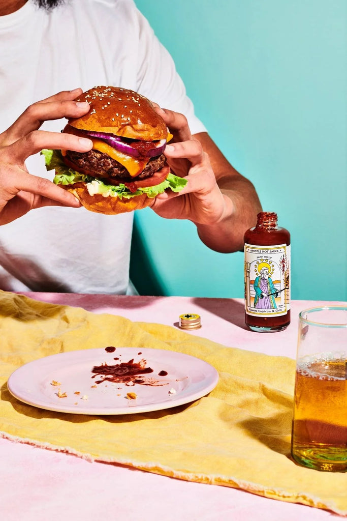 Person holding a gourmet beef burger with cheese, red onion, lettuce and Apostle Roasted Capsicum & Chilli hot sauce, with the bottle beside a messy plate and a drink on a colourful table setting