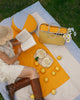 A woman in a straw hat and white dress sits on a yellow and white picnic blanket outdoors on grass, reading a book. A woven yellow picnic basket, two teacups on saucers, and scattered lemons are arranged on the blanket, with a round yellow-and-white cushion behind her.