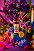Apostle Crispy Chilli Oil jar styled among vibrant purple, orange, and red flowers with a lit candle, creating a bold and colourful display scene