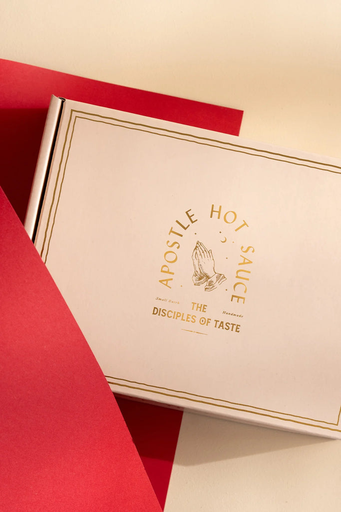Apostle Hot Sauce gift box with gold-foil logo and “The Disciples of Taste” branding, styled on a soft cream and red background.