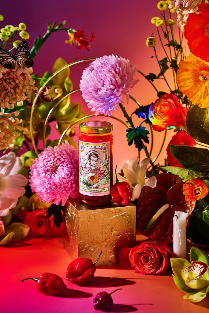 Jar of Apostle Lavender & Rosemary Hot Honey styled with vibrant florals and habanero chillies, set against a rich pink and orange backdrop