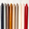 Row of nine dripless taper candles in assorted earthy tones, including black, charcoal, taupe, amber, beige, blush, ivory, and red – ideal for moody or seasonal styling.