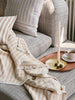 Ivory taper candle styled in a brass holder on a wooden tray beside a striped blanket and grey armchair, creating a cosy, ambient living room scene
