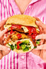 Close-up of hands holding a stacked fried chicken burger with cheese, bacon, lettuce, and Apostle Hot Sauce, against a pink striped shirt