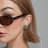 Close-up of woman wearing gold sculptural ripple earrings with tortoiseshell sunglasses and short brown hair, minimalist jewellery styling.