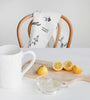 Father Rabbit tea towel with botanical sketches draped over a wooden chair, styled with a white pitcher, lemons, and citrus juicer on a wooden board.