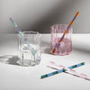 Striped glass straws in pastel and jewel tones styled in pink and clear scalloped glasses, with two straws placed diagonally on a white surface.
