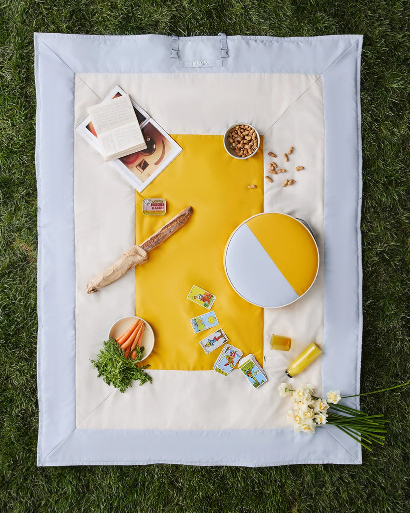A square picnic blanket laid flat on green grass, featuring a light blue border and a cream center with a large yellow geometric pattern. The blanket is styled with a book, magazine, bread, nuts, a round tray, cards, carrots, parsley, and small yellow flowers.