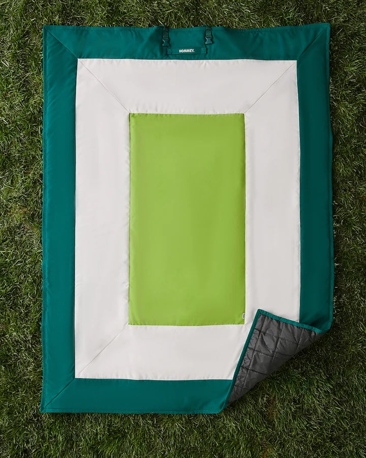 A rectangular outdoor blanket laid flat on grass, featuring a teal border, a white inner border, and a bright lime green center panel. The bottom-right corner is folded back to show a dark gray quilted underside.