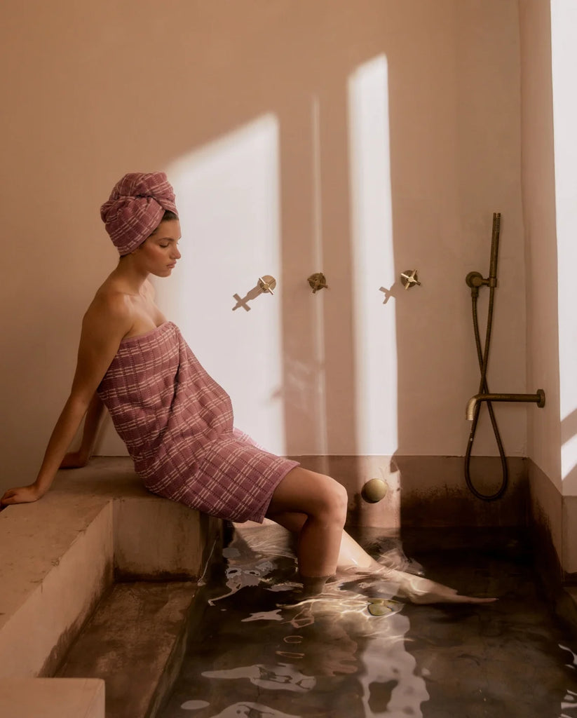 Woman sitting by a bathtub with a towel wrapped around her, sun streaming through the window.
