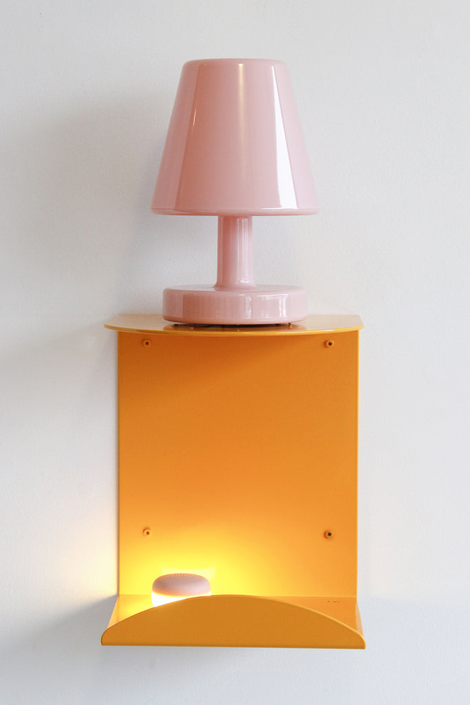 A modern wall-mounted lamp with a glossy pink tapered shade and base, mounted on a bright orange square backplate that also features an illuminated warm light below, set against a plain white wall.