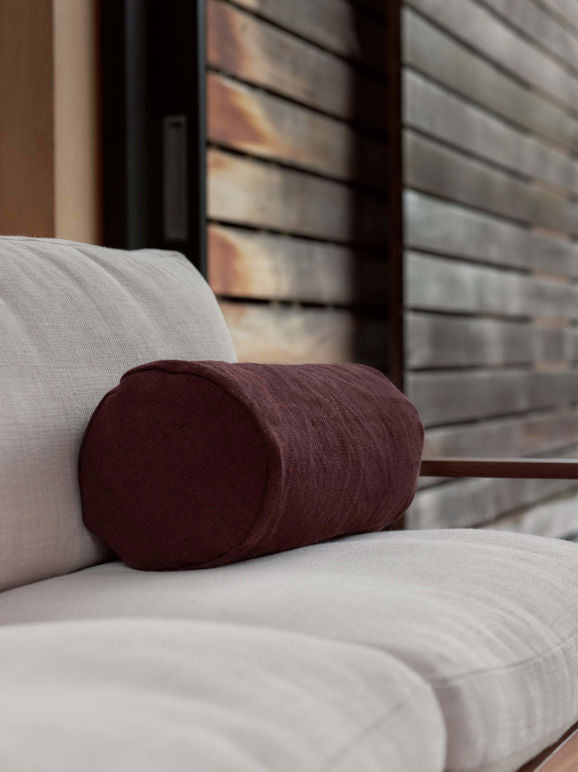 Burgundy linen bolster cushion resting on a neutral-toned outdoor sofa, styled in a modern, wooden deck setting.