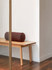 Burgundy linen bolster cushion displayed on a natural timber bench in a minimalist interior with wooden floors.