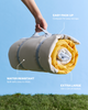 A hand holds a rolled beige and yellow travel blanket with gray straps against a blue sky background, with grass at the bottom and text callouts reading 'EASY PACK UP,' 'WATER RESISTANT,' and 'EXTRA LARGE 1.5m x 2m of blanket.