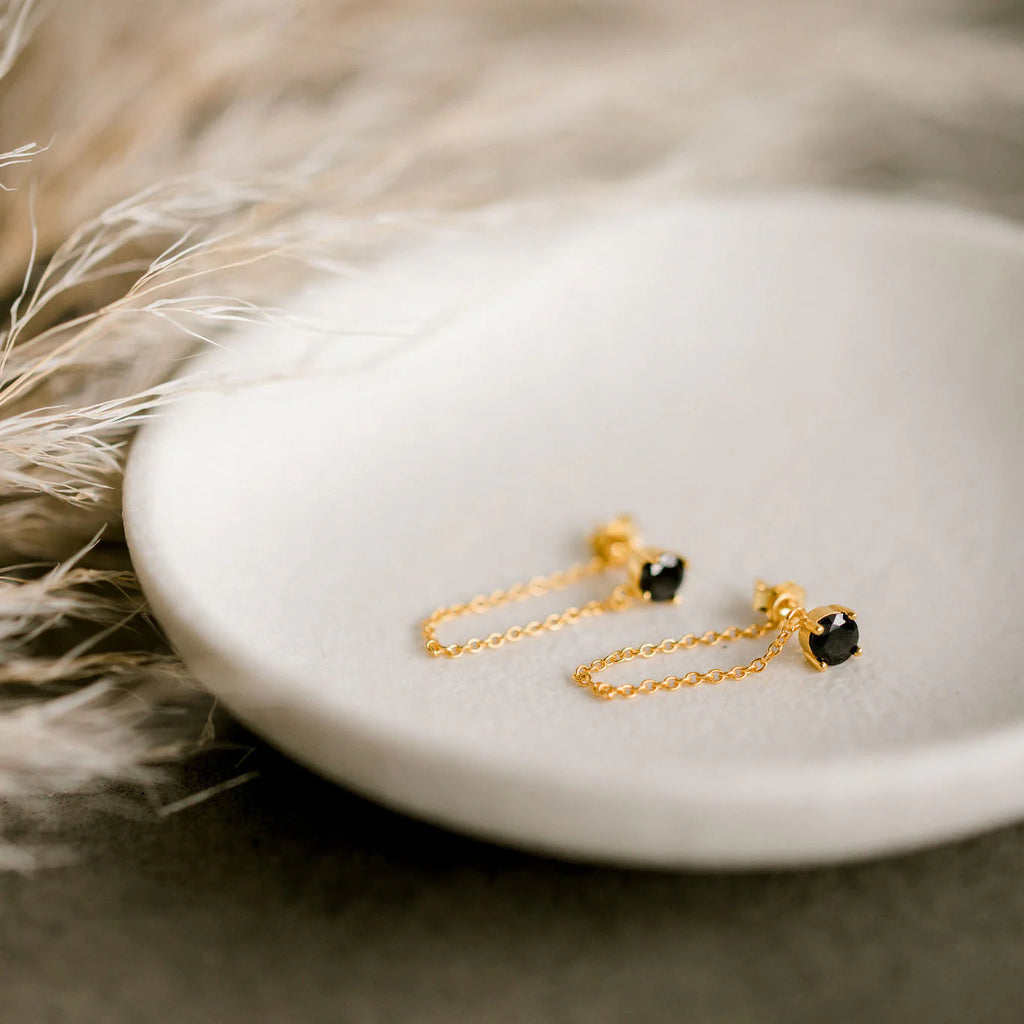 Gold chain earrings with black heart-shaped stones, styled on a ceramic dish with soft pampas grass for an elegant, minimalist presentation.