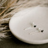 Delicate silver chain earrings with black heart-shaped stones, styled on a ceramic dish with soft, neutral-toned pampas grass in the background.