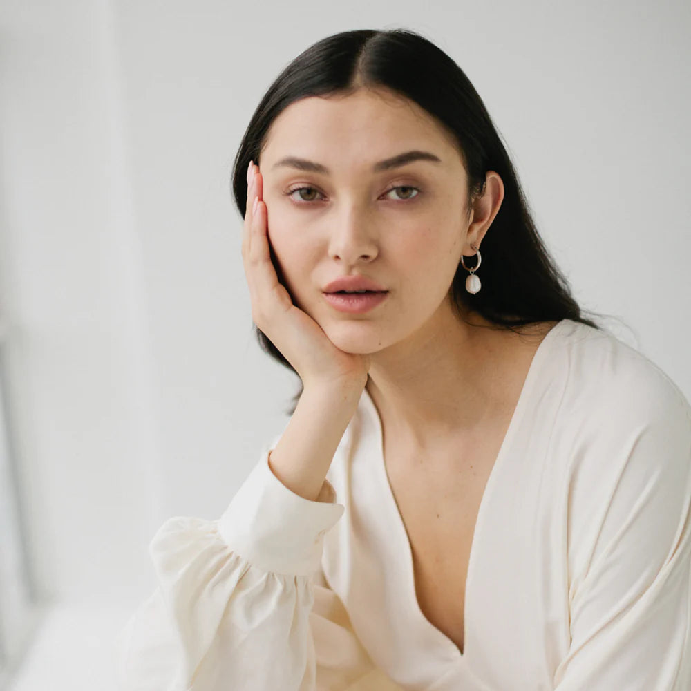 Model wearing minimalist sterling silver hoop earrings with freshwater pearl drops, styled with a soft white blouse in natural light.