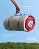 A rolled camping blanket in beige, red, and blue is being held up by a person's hand against a blue sky background, with grass at the bottom. Text callouts highlight features: 'EASY PACK UP,' 'WATER RESISTANT,' and 'EXTRA LARGE 1.