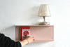A minimalist wall-mounted shelf in a dusty pink color, styled with a clear glass table lamp and a red book, with a person's hand holding a white mug in the foreground against a white wall.