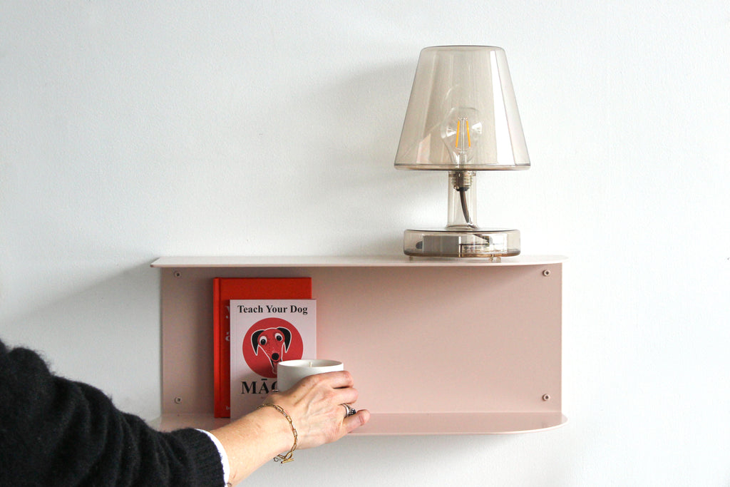 A minimalist wall-mounted shelf in a dusty pink color, styled with a clear glass table lamp and a red book, with a person's hand holding a white mug in the foreground against a white wall.