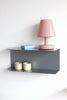 A modern dark gray metal wall shelf mounted on a white wall, styled with a pink table lamp, a light blue box, and two cream ceramic cups.