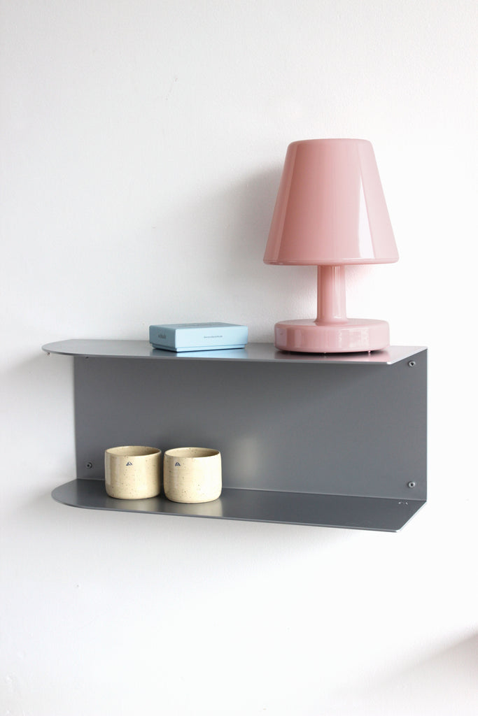 A modern dark gray metal wall shelf mounted on a white wall, styled with a pink table lamp, a light blue box, and two cream ceramic cups.