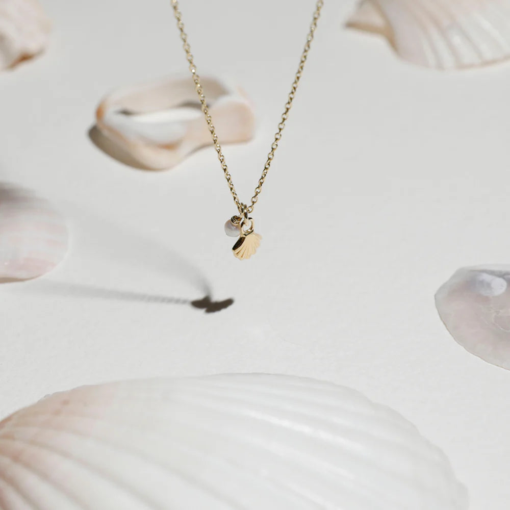 Gold necklace with a shell charm and pearl pendant, styled among natural seashells on a light background for a summery, coastal-inspired aesthetic.