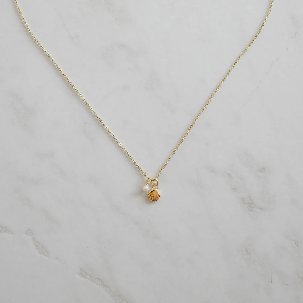 Delicate gold necklace featuring a small shell charm and pearl pendant, styled on a marble surface for a coastal-inspired, minimalist look.