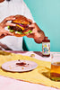 Person holding a gourmet beef burger with cheese, red onion, lettuce and Apostle Roasted Capsicum & Chilli hot sauce, with the bottle beside a messy plate and a drink on a colourful table setting