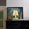 Framed abstract painting with a lamp on top of books against a white wall.