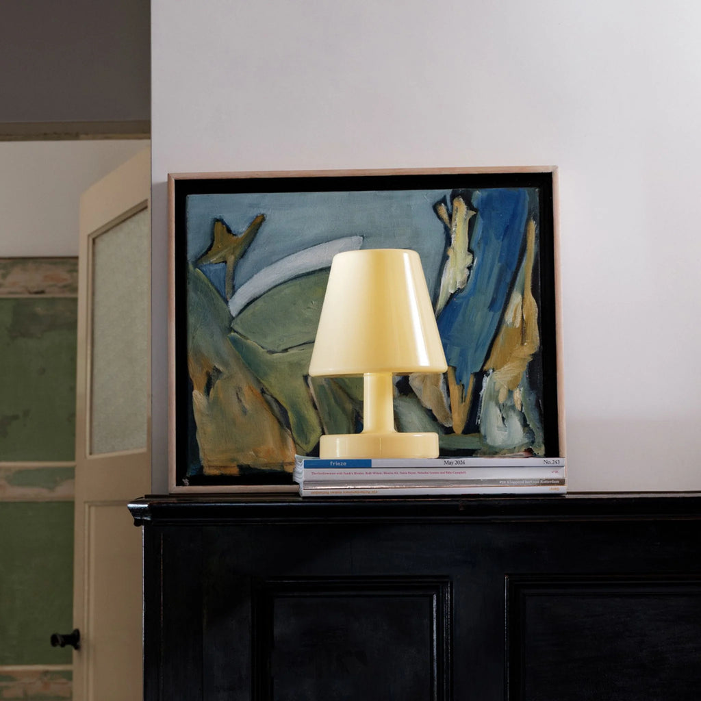 Framed abstract painting with a lamp on top of books against a white wall.