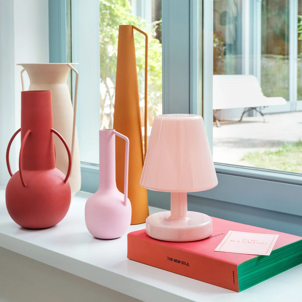 A pink table lamp sits on a stack of colorful books on a windowsill, with several decorative vases and a tall orange object in the background near a bright window.