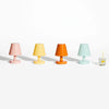 Four small table lamps in pastel pink, yellow, orange, and mint green are lined up on a white background, with a small glass of lemonade and a yellow-and-white striped straw placed to the right for scale.