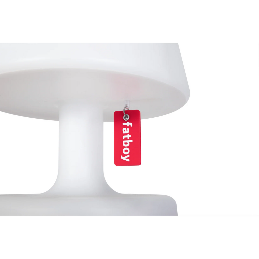 A red rectangular acrylic tag with the brand name 'fatboy' printed in white, hanging from a small metal ring under the edge of a white pedestal lamp.
