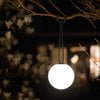 A spherical outdoor lantern hanging from a tree branch at night, glowing with a bright white light against a dark blurred background.