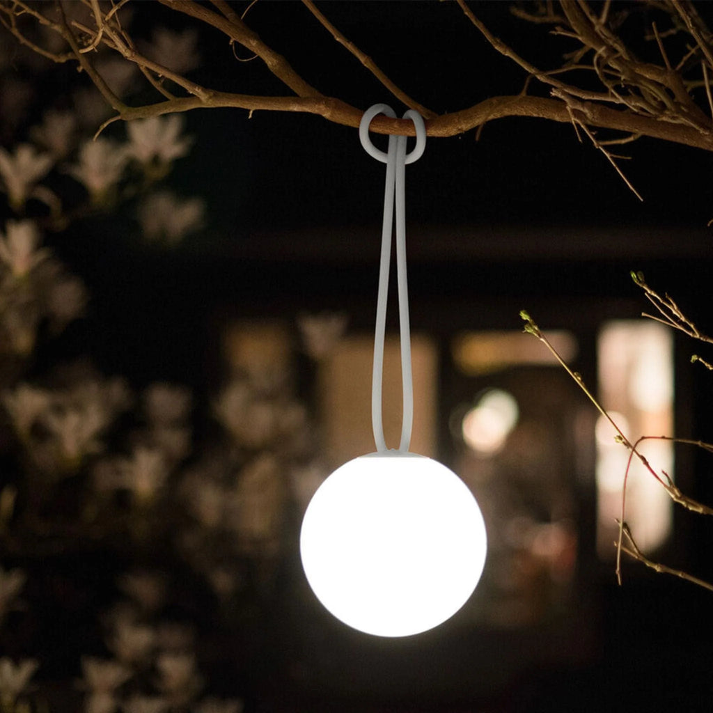 A spherical white hanging pendant light suspended from a tree branch at night, glowing brightly against a dark blurred outdoor background.