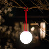A spherical white hanging lantern with a bright red hanging cord, suspended from a tree branch at night against a dark, softly blurred outdoor background.