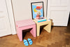 A pink upholstered stool sits beside a pale yellow side table in a room with white paneled doors and a wood parquet floor. A framed print, books, a small cup, and blue slide sandals are placed on and around the furniture.
