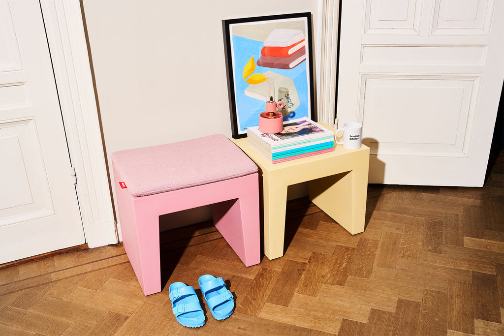 A pink upholstered stool sits beside a pale yellow side table in a room with white paneled doors and a wood parquet floor. A framed print, books, a small cup, and blue slide sandals are placed on and around the furniture.