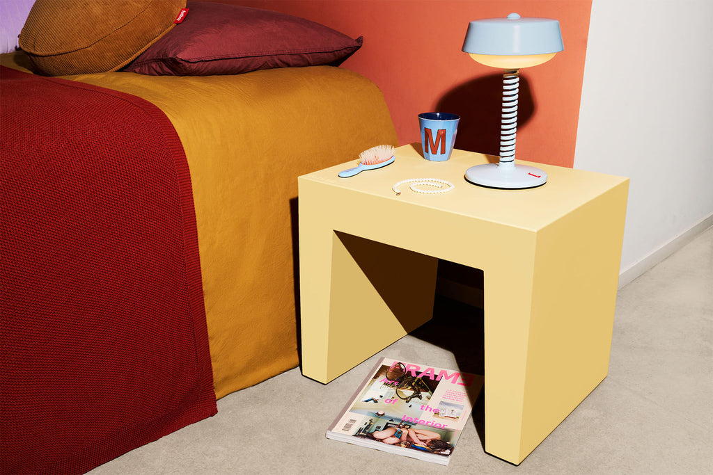 Yellow side table next to a bed with a lamp, cup, and magazine.