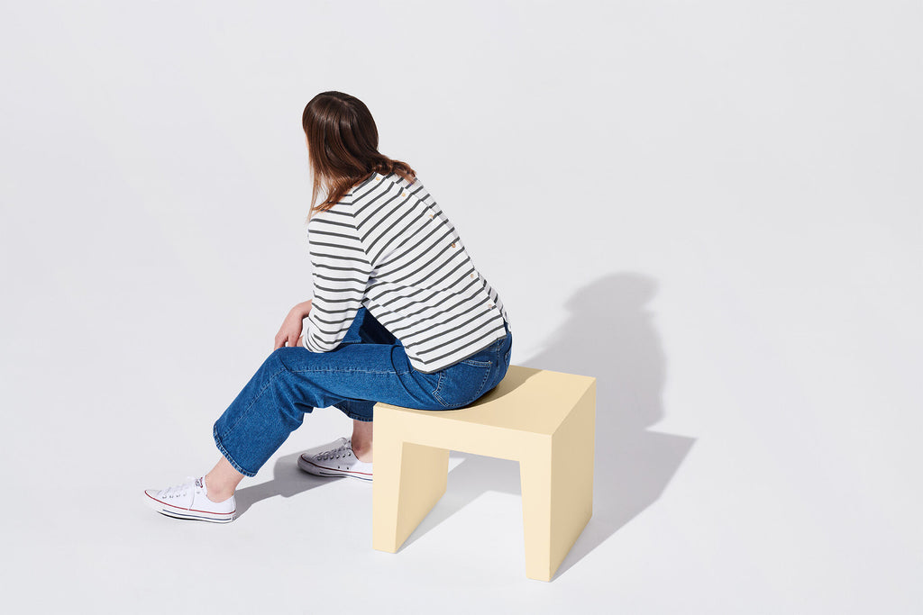 A woman sits on a pale yellow block stool, photographed from behind against a clean white studio background. She wears a black-and-white striped long-sleeve top, blue jeans, and white sneakers.