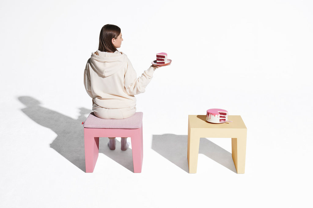 A woman in a cream hoodie sits on a pink block stool, holding a pink cake. A small light wood table to the right holds a matching pink cake.