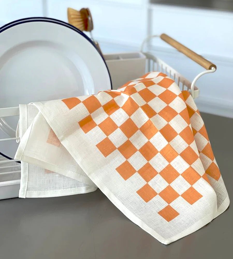 Orange and white checkerboard tea towel draped over a white dish rack, beside stacked white plates in a kitchen setting.