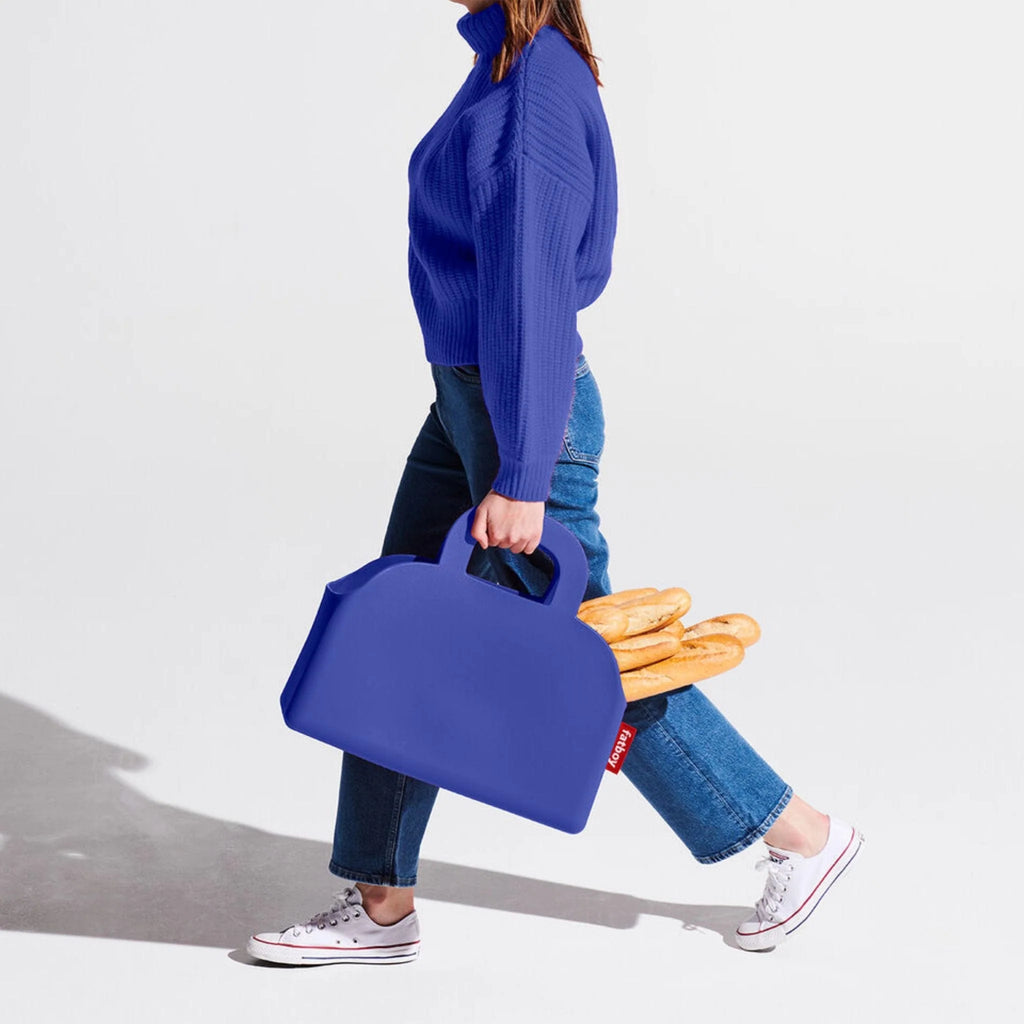 A woman in a blue sweater, jeans, and white sneakers carries a large royal blue silicone bag filled with baguettes, walking 